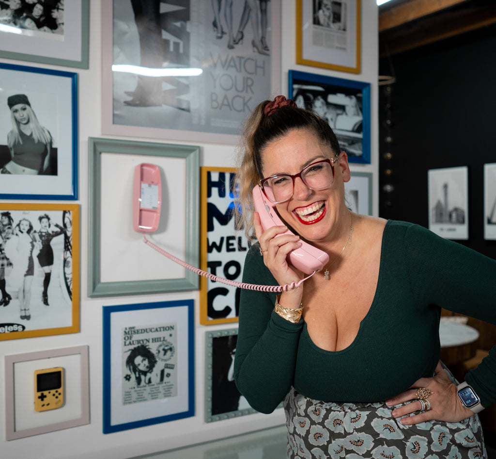 A person smiling and holding a pink telephone in front of a wall with framed photographs and artwork.