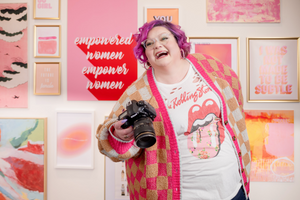 A person with pink hair and a plaid shirt holding a camera, standing in front of a wall with framed artworks and text that reads 'empowered women empower women'.