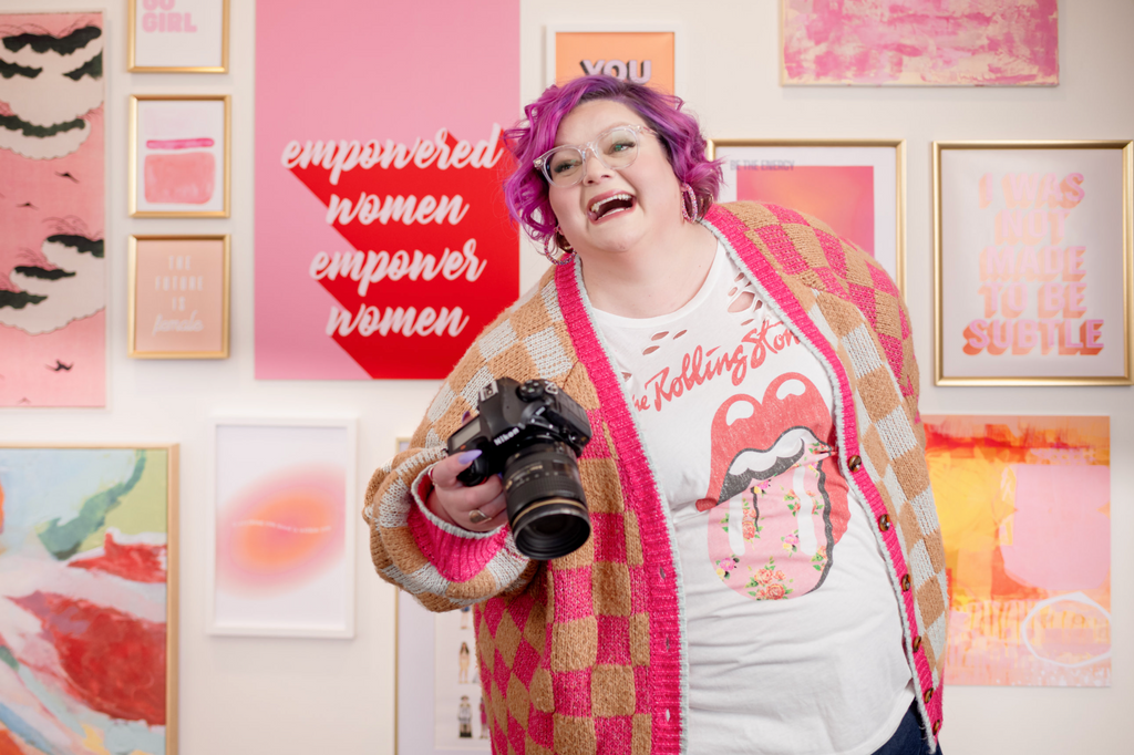 A person with pink hair and a plaid shirt holding a camera, standing in front of a wall with framed artworks and text that reads 'empowered women empower women'.