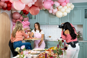 An image showing a group of people at a party or gathering, with balloons and food in the background. It appears to be a festive atmosphere.